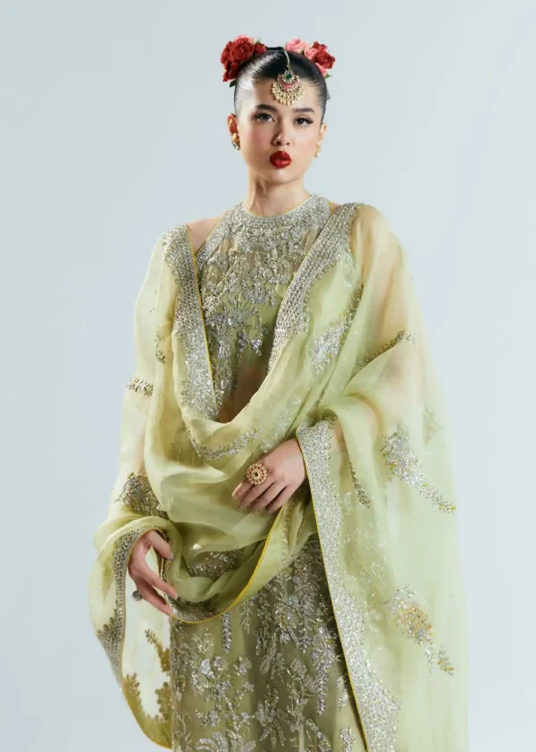 A model in a heavily embroidered pastel green traditional outfit with silver embellishments, styled with bold red lips, floral accessories, and ethnic jewelry.