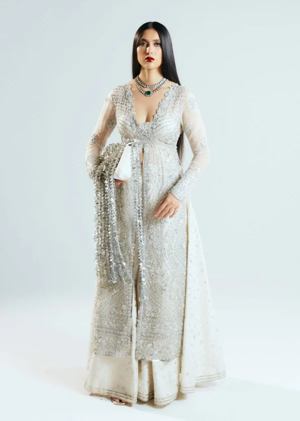 A model in an ivory designer outfit with intricate silver embellishments, sheer sleeves, and a floor-length silhouette.